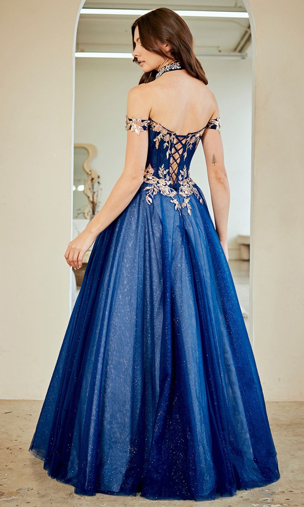 A woman in the Amelia Couture 8063 long prom dress, an off-the-shoulder dark blue gown with sequin appliquรฉ, floral embroidery, and a lace-up back, stands before a mirror.